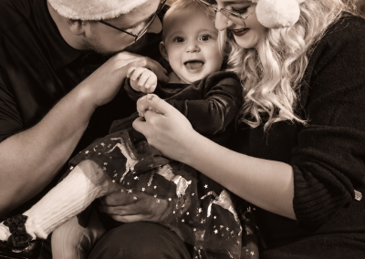 Black-and-white Christmas family portrait of two parents in Santa hats cuddling their smiling baby in front of a softly lit Christmas tree.