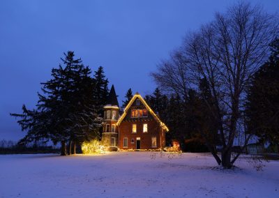 Historic Drummond Manor in Waterdown, Ontario, decorated with glowing Christmas lights on a snowy winter evening, surrounded by tall trees beneath a deep blue twilight sky.