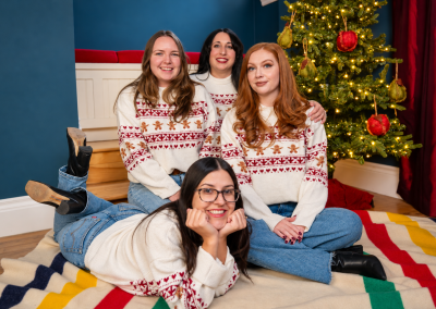 Four women in matching Christmas sweaters posing beside a decorated tree at Drummond Manor in Waterdown